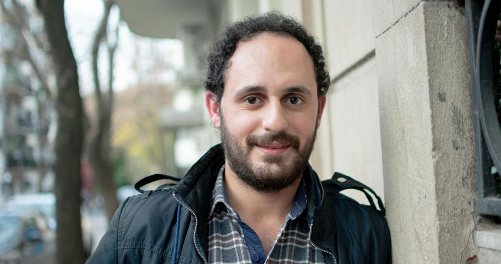 A young man leaning against a wall in a jacket, smiling kindly