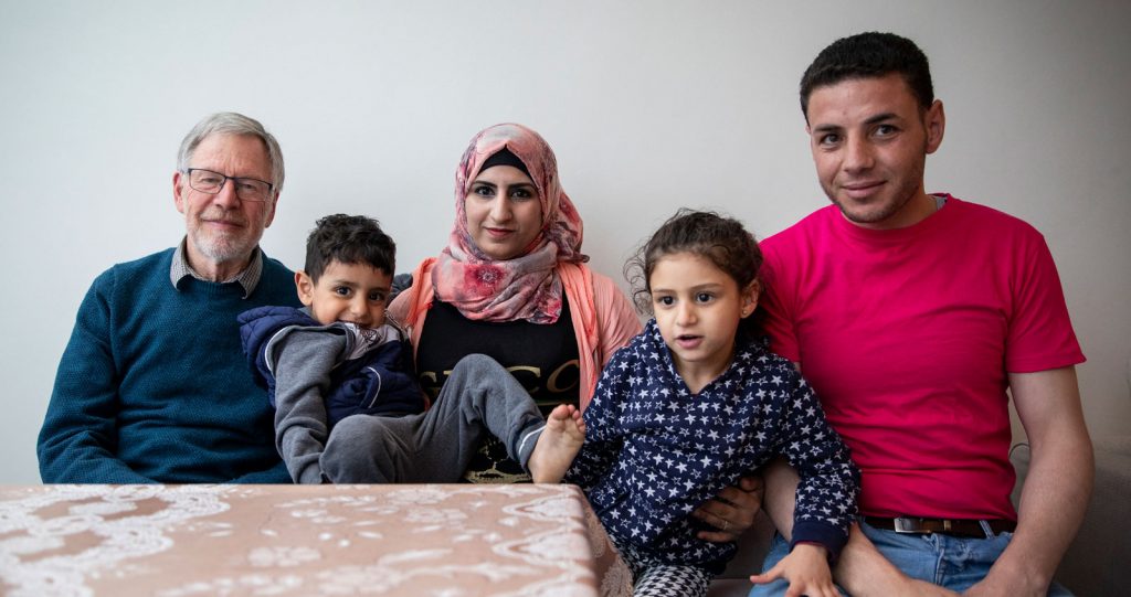 A family and an elderly man sit around a kitchen table. A young couple have their two children perched in their laps.