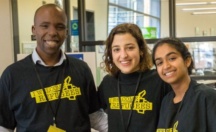 Three young people wearing black and yellow 'I welcome refugees' tshirts, smiling warmly