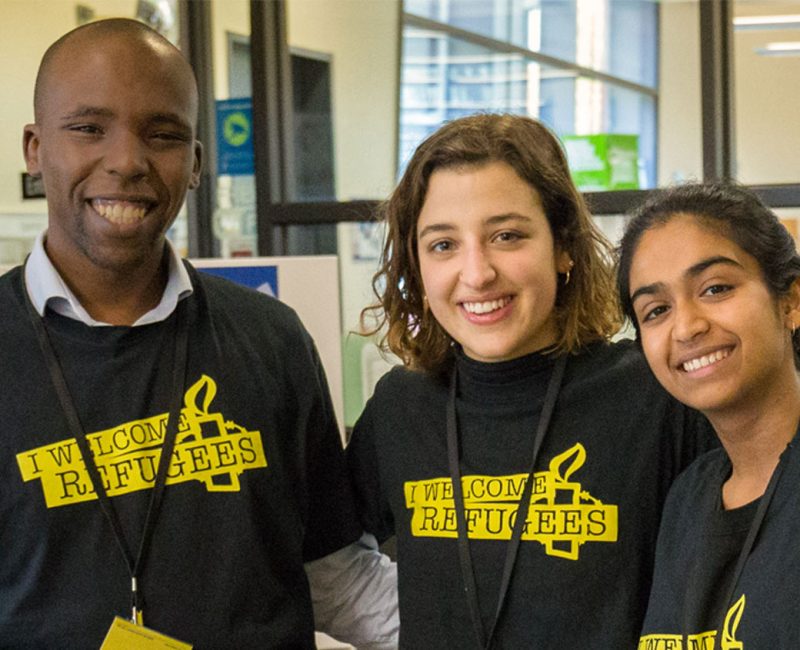 Three young people wearing black and yellow 'I welcome refugees' tshirts, smiling warmly