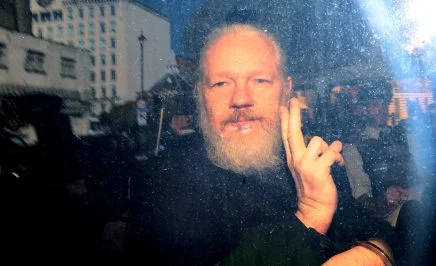 Julian Assange, a man with a white beard, raises his hand in a peace gesture with media and police around him. Twilight evening, bright flash of light to the right.