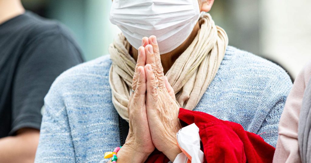 A person wears a white facemask, blue sweater and beige scarf. Their hands are doing a prayer.