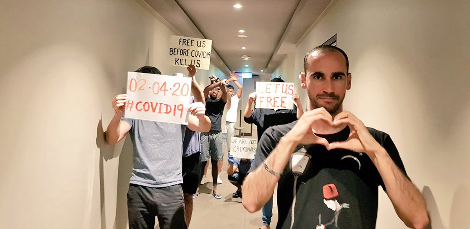 A small group of men stand in a hotel hallway, holding handmade signs asking 'Free us before COVID19 kills us'. One man creates heart symbol with his hands.