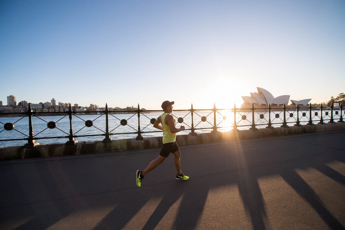 A man jogs along the harbour in Sydney, the sun low on the horizon and the Sydney Opera House in the background.