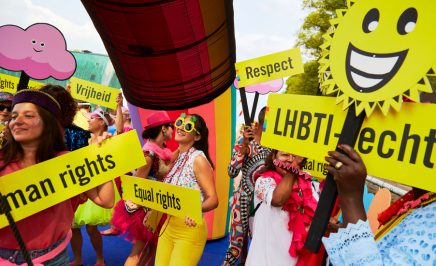 A very colourful rainbow parade of people dancing, with fun yellow signs saying human rights, equal rights, respect & LGBTI. One person holds a smiling sun placard.