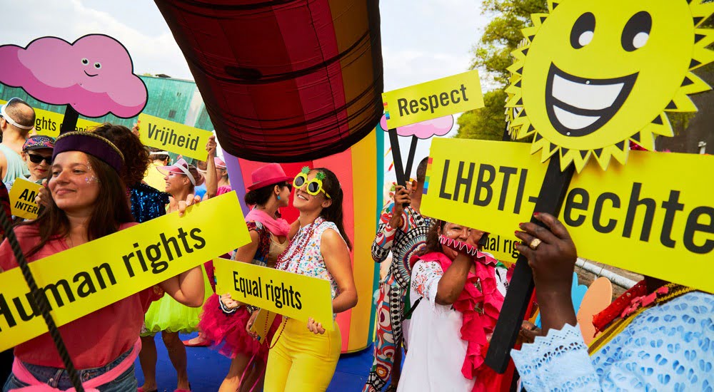 A very colourful rainbow parade of people dancing, with fun yellow signs saying human rights, equal rights, respect & LGBTI. One person holds a smiling sun placard.