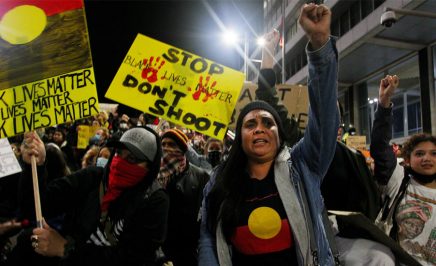 Protestors chant and bend down on their knees. They hold handmade red, yellow and black signs saying 'stop don't shoot' and Black Lives Matter. Woman in front raises her fist and is wearing an Aboriginal flag tshirt.