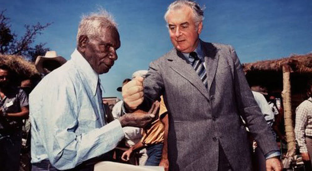 Vincent Lingiari (left) holds out his palm. Gough Whitlam (right) holds his hand above Vincent's and pours red earth into Vincent's palm. Brilliant blue sky above them.