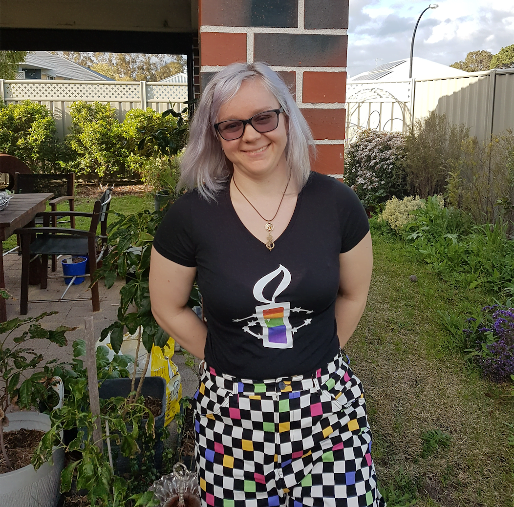 Image of LGBTIQ activist Hannah Wahlsten, smiling at standing in her yard. She's wearing rainbow-coloured checkered pants, an Amnesty tshirt with a rainbow candle, and black glasses.
