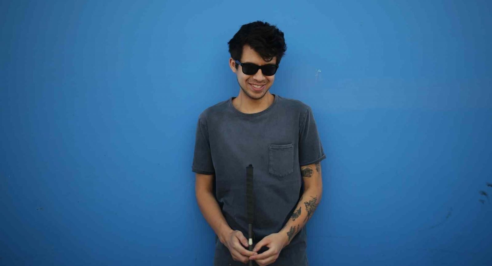 Gustavo stands with a can and dark sunglasses in a grey shirt before a blue background.