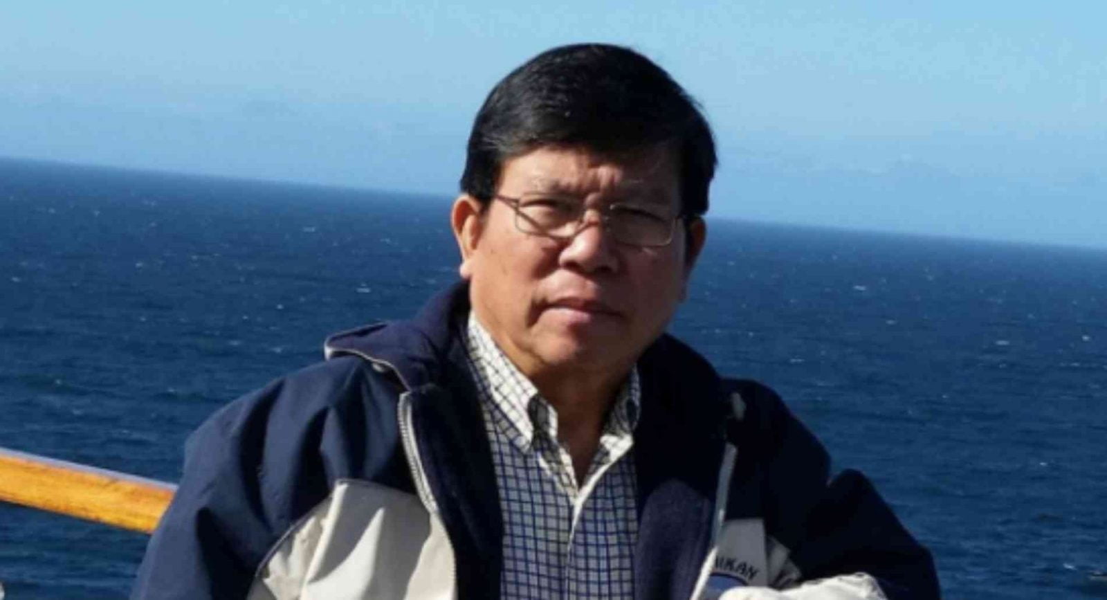 Mr Chau wears a jacket and glasses, and stands in front of the ocean.