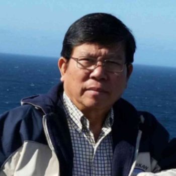 Mr Chau wears a jacket and glasses, and stands in front of the ocean.