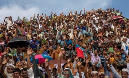 A crowd of Rohingya people, of all ages, sit and stand in a crowd with their hands raised.
