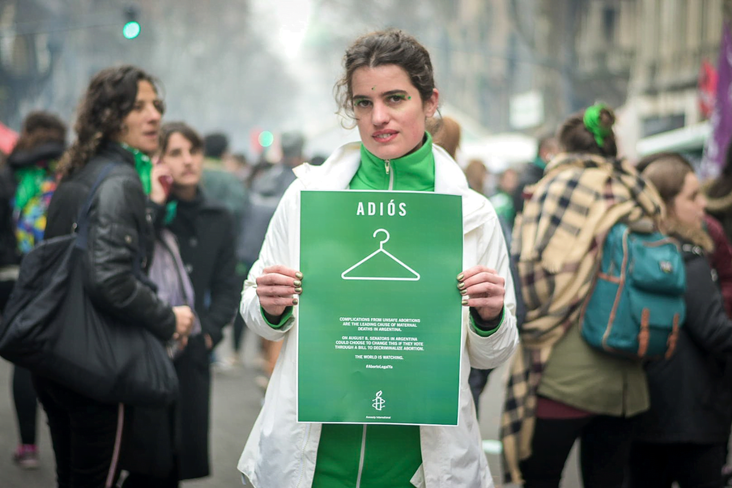 A woman wearing green and white holds a sign at an Argentinian protest calling for reproductive rights. The sign reads 'Adios' and has an image of a coathanger.