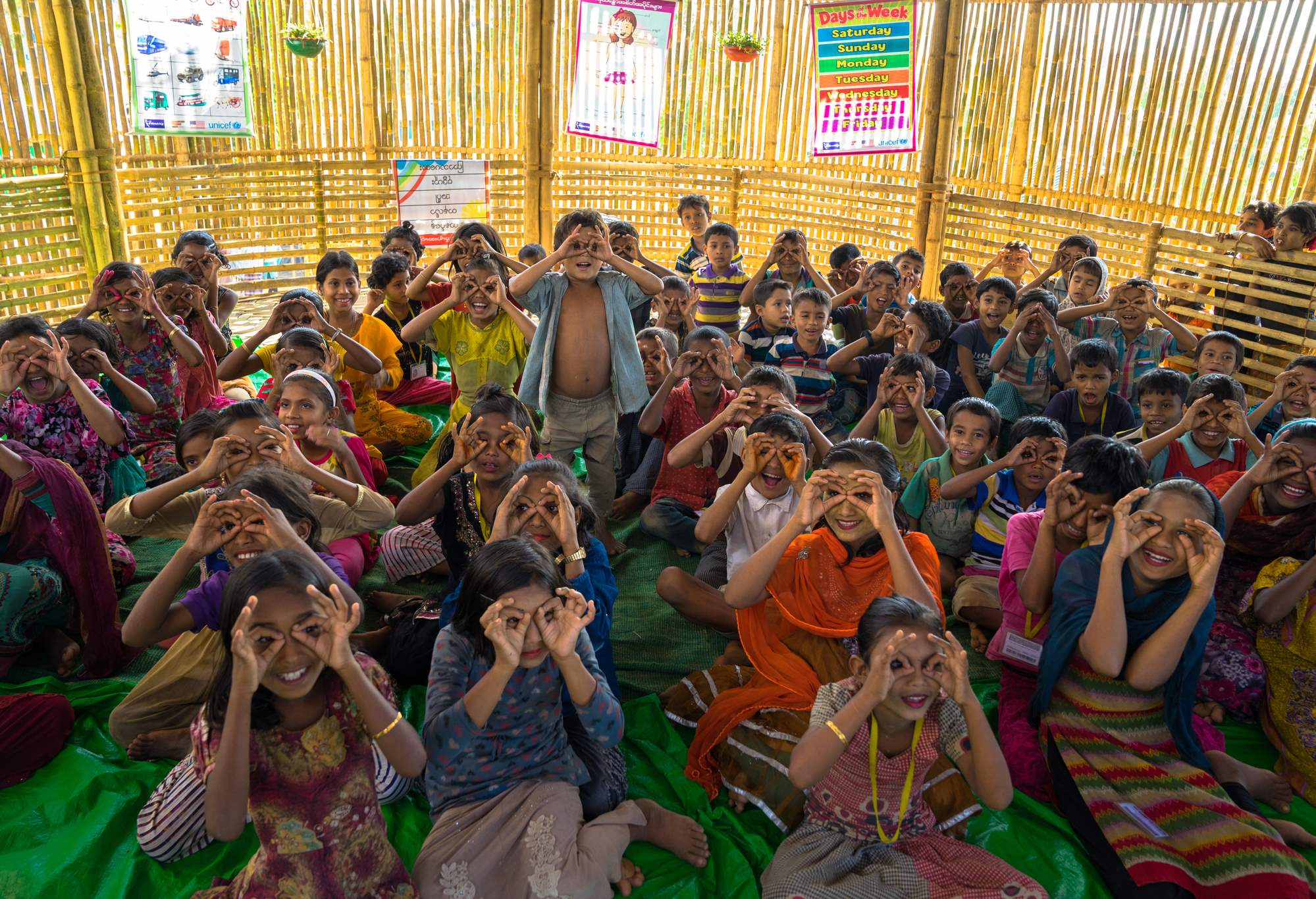 A room of Rohingya children sitting on the classroom floor smiling, holding up their fingers which form circles around their eyes.