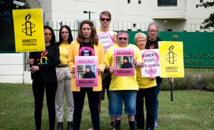 A group of activists in yellow shirts hold pink posters with a photo of Nassima and the words 