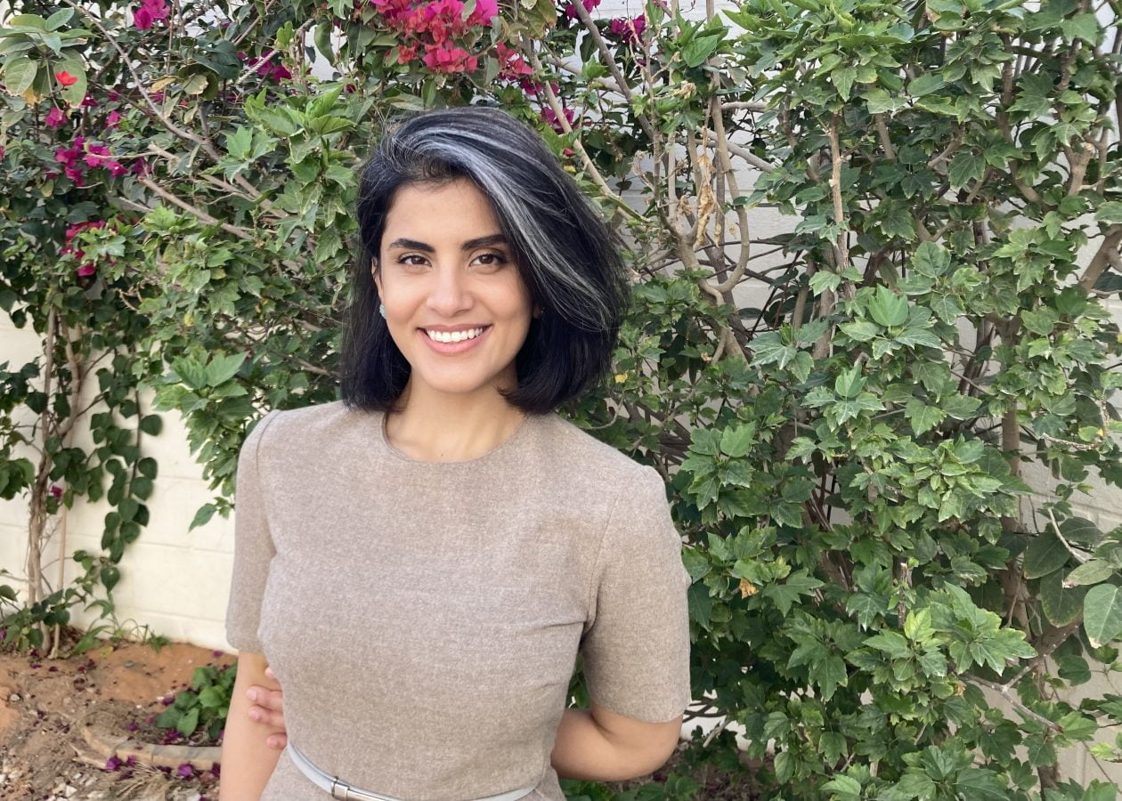 Loujain with black hair and a gray dress stands in front of a vine.
