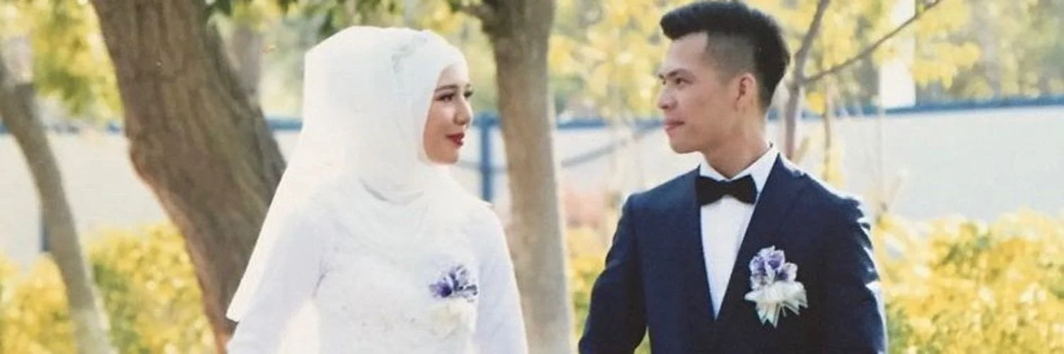 Mehray and Mirzat on their wedding day. Mehray wears a white wedding dress and hijab, Mirzat wears a black blazer. They are looking at each other.