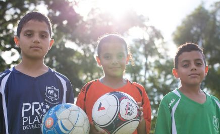 Three children holding soccer balls and wearing soccer jerseys in a sunlit park.