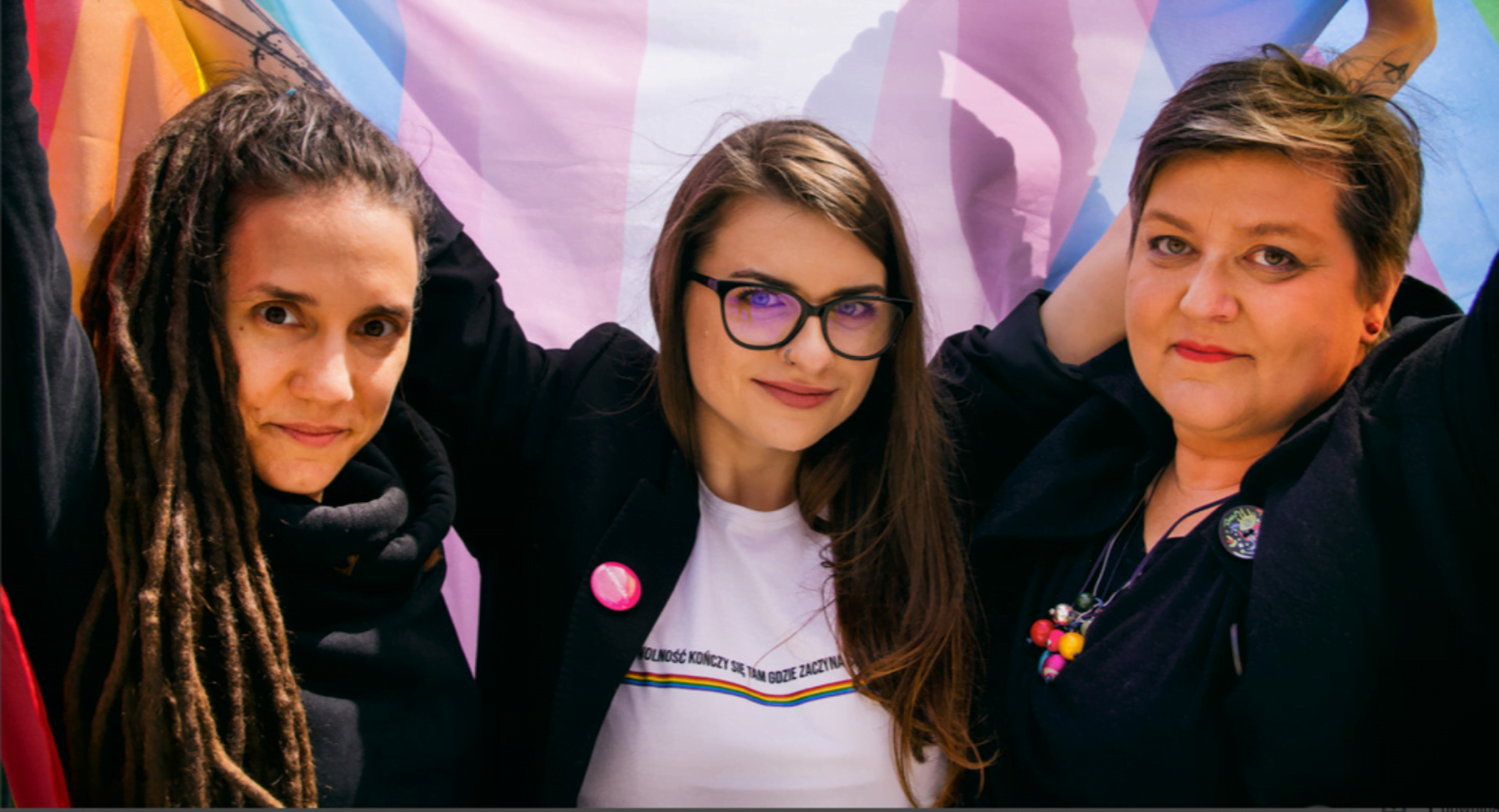 Elżbieta, Anna and Joanna stand in front of an LGBTQIA+ flag