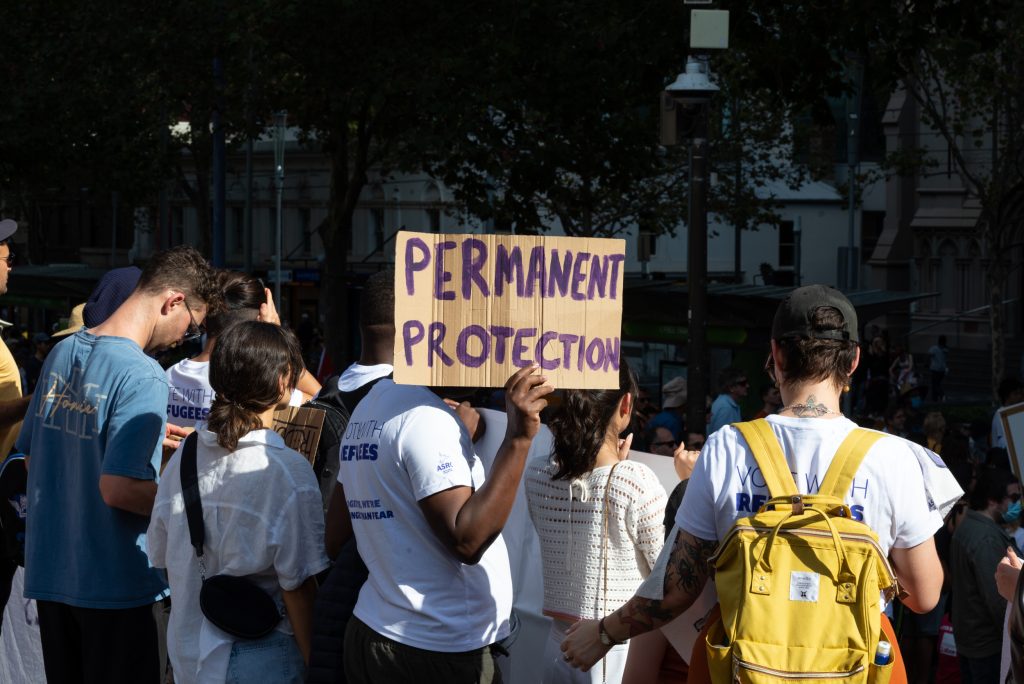 A man stands in a protesting crowd for refugee rights, holding a sign that says 'Permanent Protection'