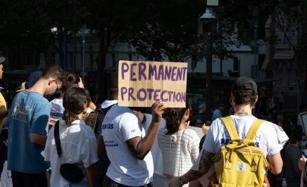 A man stands in a protesting crowd for refugee rights, holding a sign that says 'Permanent Protection'