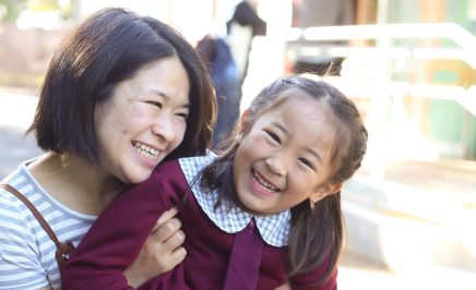 Mother holds daughter. Both are smiling. Daughter is in school uniform.