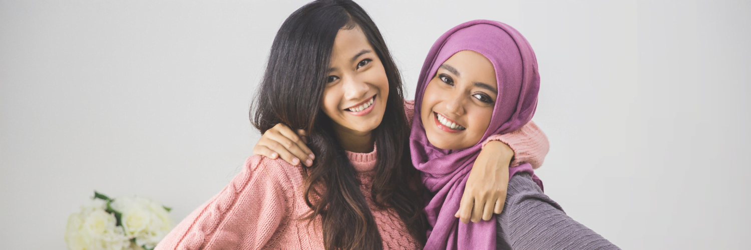 Two young women of colour, smiling. The girl on the left has long dark hair and has her arm around the girl on the right, who is wearing a purple hijab.