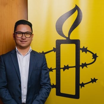 Amnesty International Australia Refugee Rights Campaigner Zaki Haidari pictured next to an Amnesty logo of a candle surrounded by barbed wire on a yellow background.
