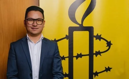 Amnesty International Australia Refugee Rights Campaigner Zaki Haidari pictured next to an Amnesty logo of a candle surrounded by barbed wire on a yellow background.