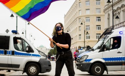 Protestor standing in between two police cars waving LGBTQ+ flag.