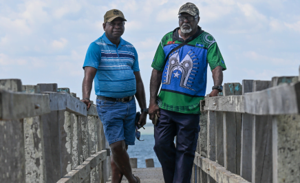 Two First Nations traditional owners, Uncle Pasul and Uncle Pabai, stand on a dock in front of the ocean in the Torres Strait Islands