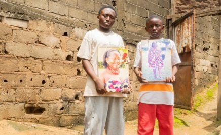Two young boys (Dorgelesse's son and nephew) hold images of Dorgelesse outside their family home in Cameroon