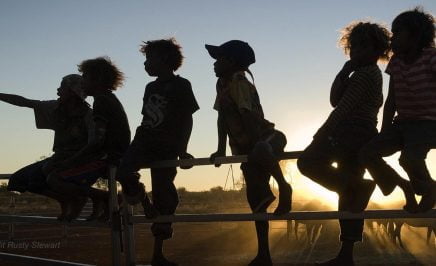 Indigenous children are silhouetted against the sunset as they sit on a fence