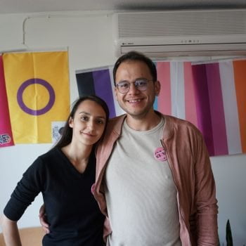 Melike Balkan and Özgür Gür smile at the camera, with an arm around each other, standing in front of a wall with Pride flags hanging on it