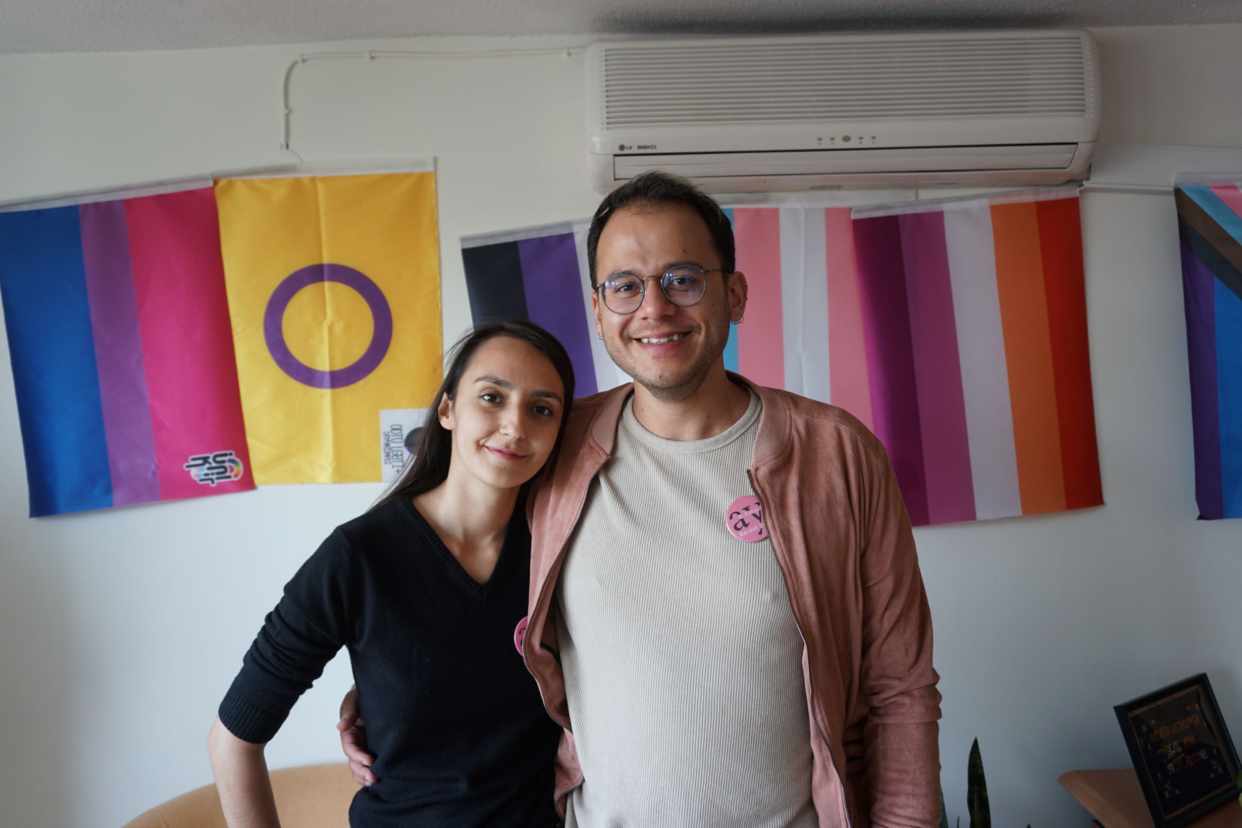 Melike Balkan and Özgür Gür smile at the camera, with an arm around each other, standing in front of a wall with Pride flags hanging on it