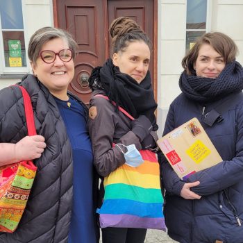 Elżbieta, Anna and Joanna stand on the street, smiling at the camera, holding rainbow tote bags