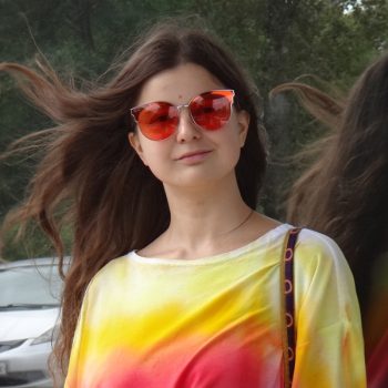 Yulia Tsvetkova smiling, wearing red sunglasses and a rainbow long sleeve top.