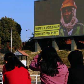 A group of young women in Qatar look at a billboard of a migrant worker with text 'What's a world cup without fair play?'