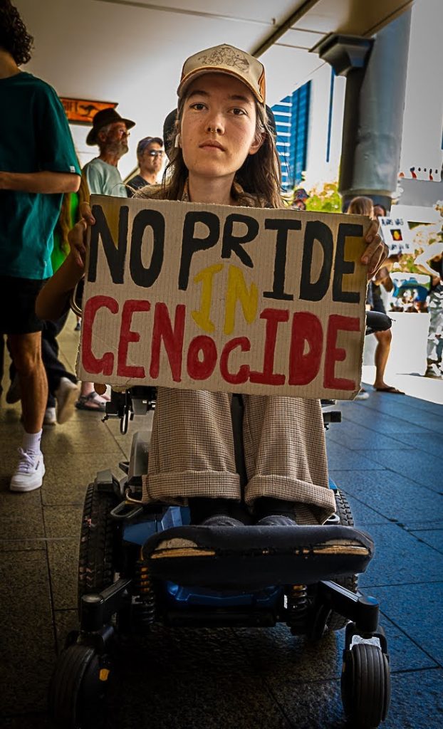 A protestors in a wheelchair at Invasion Day 2024 rally in Perth holds a sign with the words 'No Pride in Genocide'