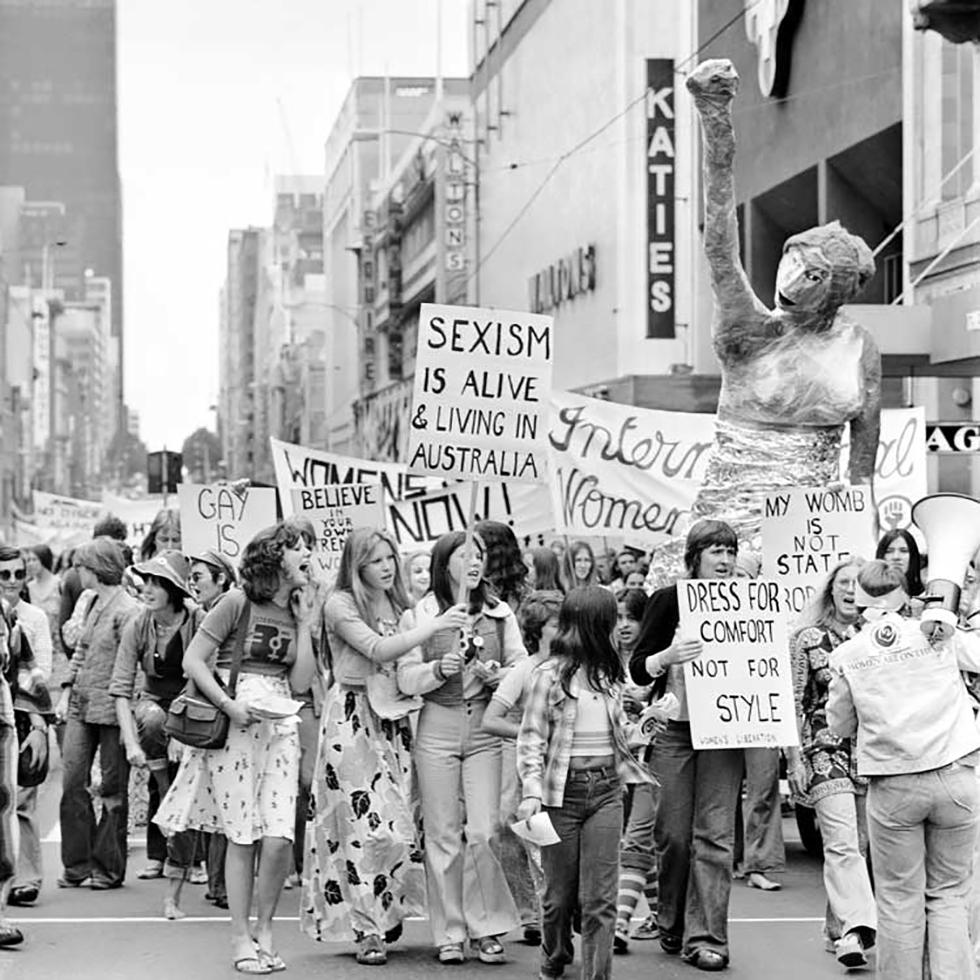 A black and white image of women protesting with handheld signs at the first International Women's Day rally in Australia. Signs read "dress for comfort not style" and "sexism is alive and living in Australia".