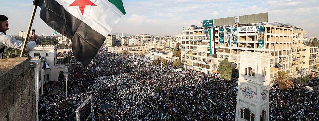 TOPSHOT - A Syrian flag flutters above crowds gathering during celebrations marking one year since a lightning Islamist-led offensive that eventually toppled the country's longtime ruler, in central Hama on December 5, 2025. (Photo by OMAR HAJ KADOUR / AFP via Getty Images)